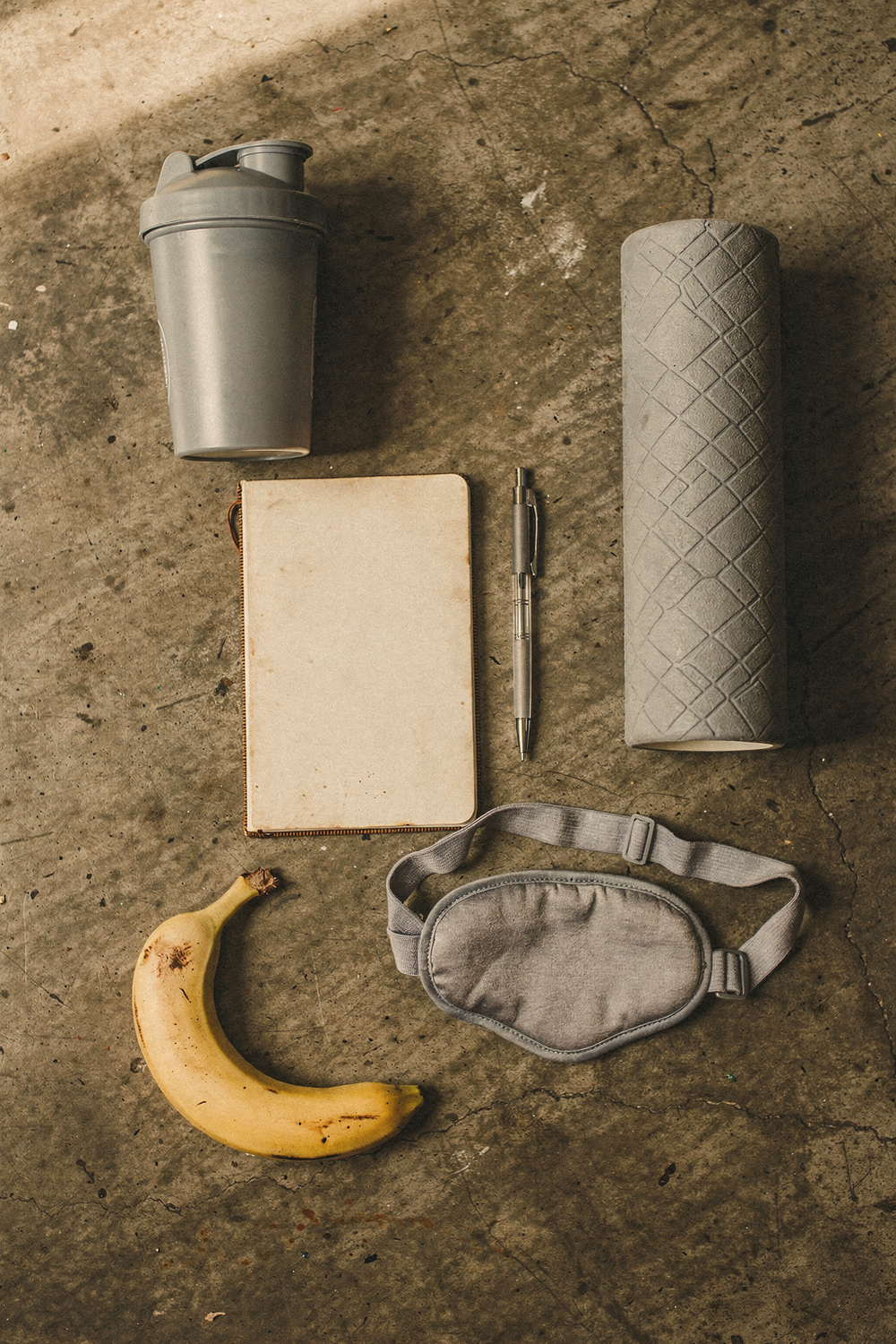 A protein shaker, a training journal with pen, a foam roller, a sleep mask, and a banana arranged in a loose grid. , no faces, no hands