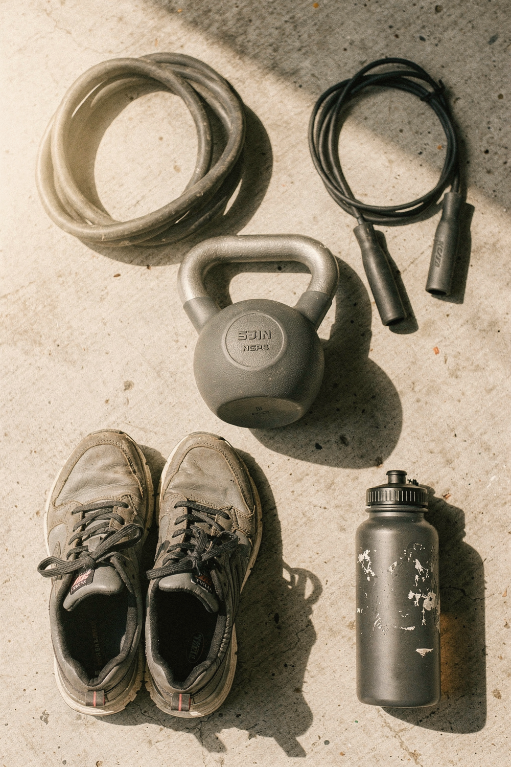 A resistance band coiled loosely, a jump rope, a kettlebell, a pair of trainers, and a water bottle arranged in a loose grid. , no text, no logos