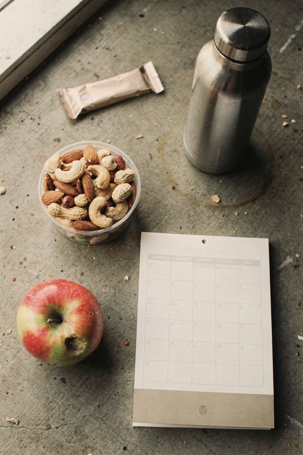 A vending machine candy bar next to a small container of mixed nuts and an apple, a desk calendar showing the current week, a reusable water bottle. Practical, real, zero-judgment. , no faces, no hands
