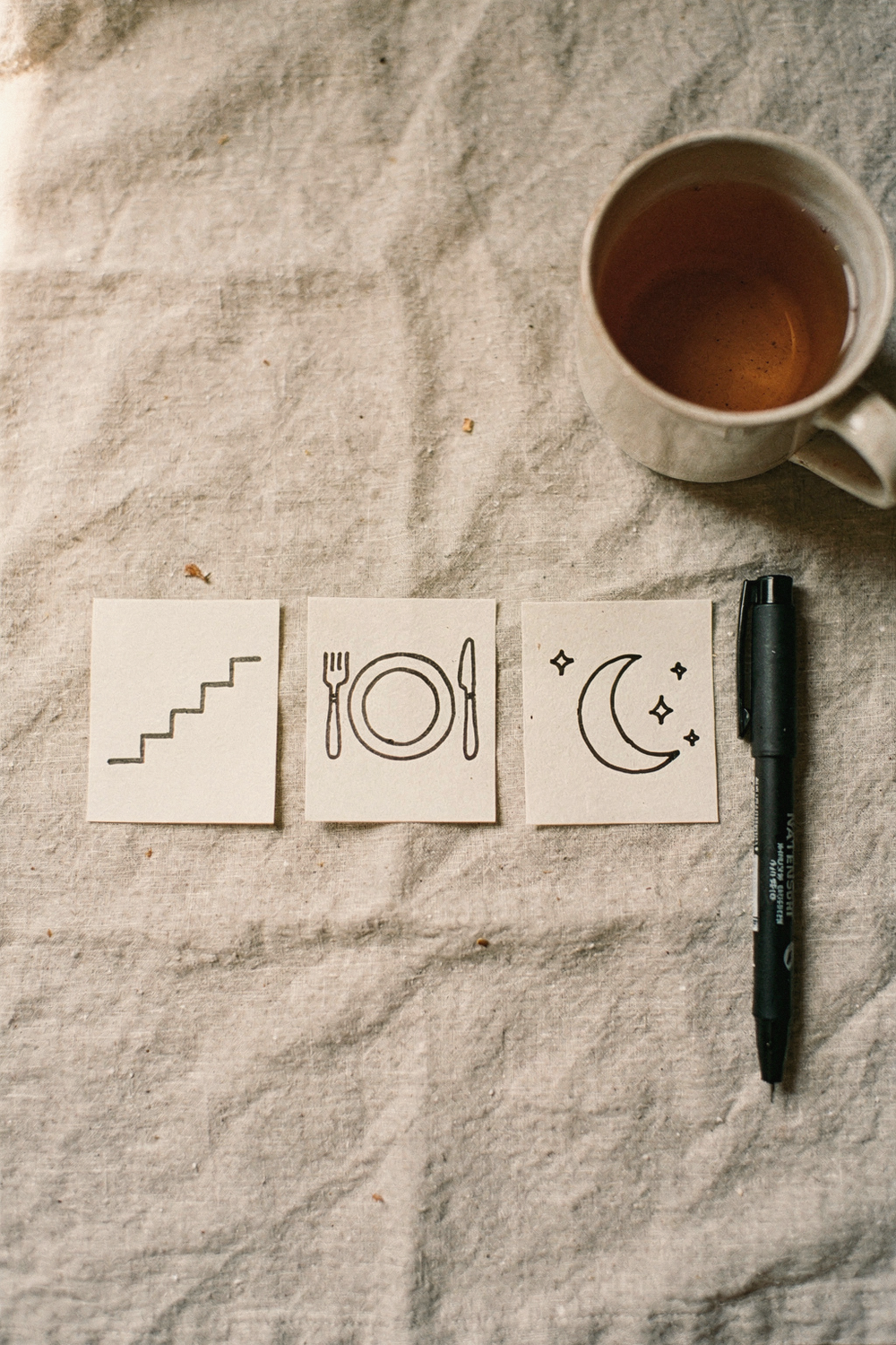 Three small index cards laid in a row, each marked with a simple hand-drawn icon for steps, food, and sleep, beside a felt-tip pen and a ceramic mug of tea. , no text, no logos