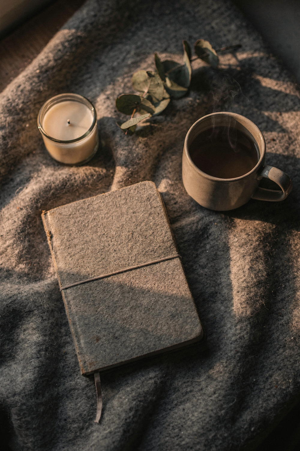 A small reflection journal closed with an elastic band, a steaming mug, a candle unlit, a sprig of eucalyptus. Restful, honest, human. , no text, no logos