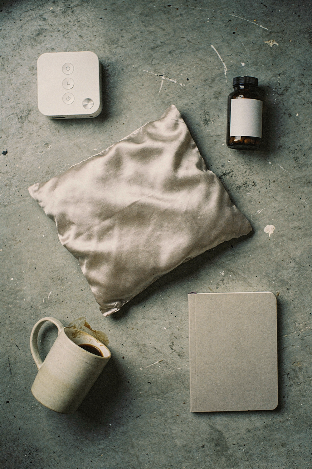 A fancy silk pillowcase, a white noise machine, a bottle of magnesium supplements, a coffee mug tipped on its side, a small notebook. , no faces, no hands