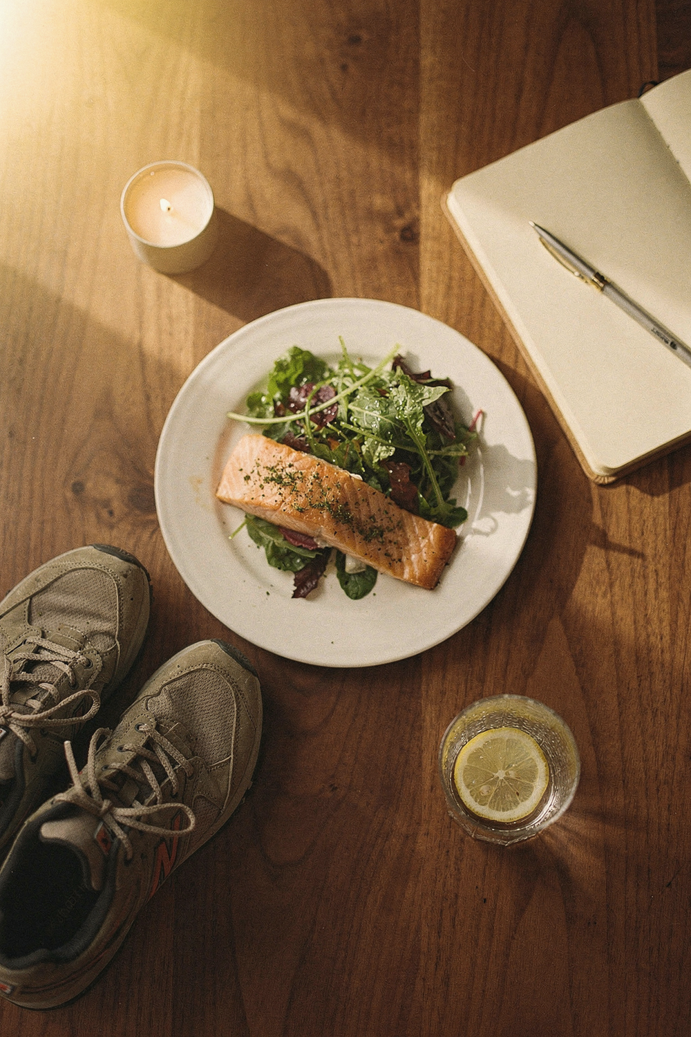 A dinner plate with salmon and greens, a pair of walking shoes, a journal with a pen, a small candle, a glass of water with lemon. Holistic, grounded, purposeful. , no faces, no hands