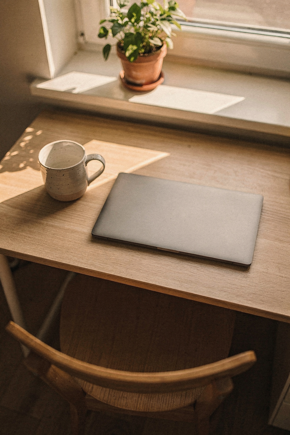 A wooden chair pushed back from a desk, a closed laptop, an empty ceramic mug, and a small potted plant on the windowsill behind. Quiet, uncluttered, inviting. , no text, no logos