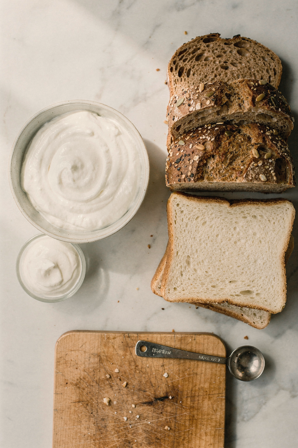 A bowl of Greek yogurt beside a small container of sour cream, whole grain bread next to white bread, a measuring spoon, and a small cutting board