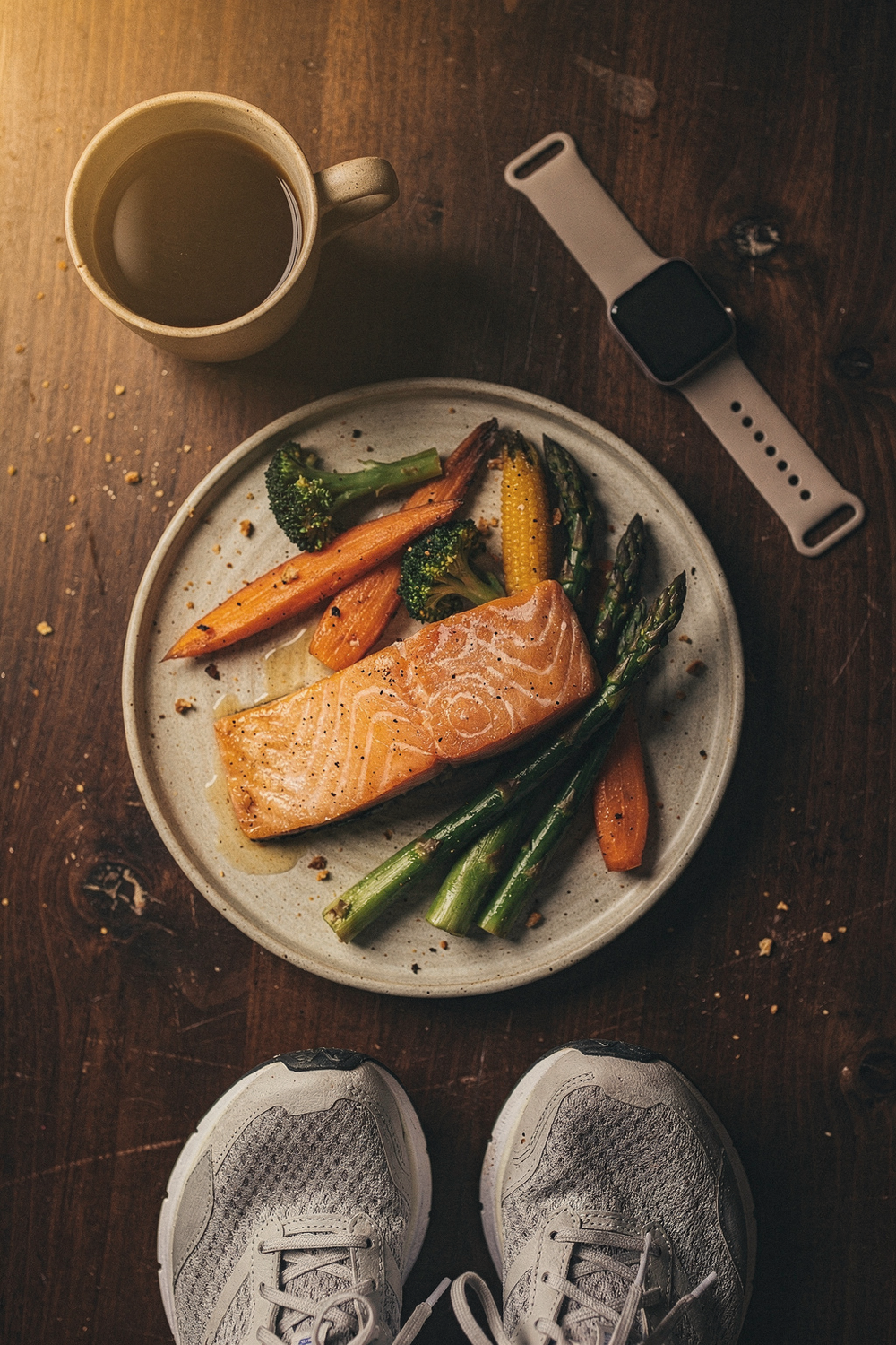 A plate with salmon and vegetables, a coffee cup, a sleep tracker watch, and a pair of running shoes at the edge. Grounded, honest