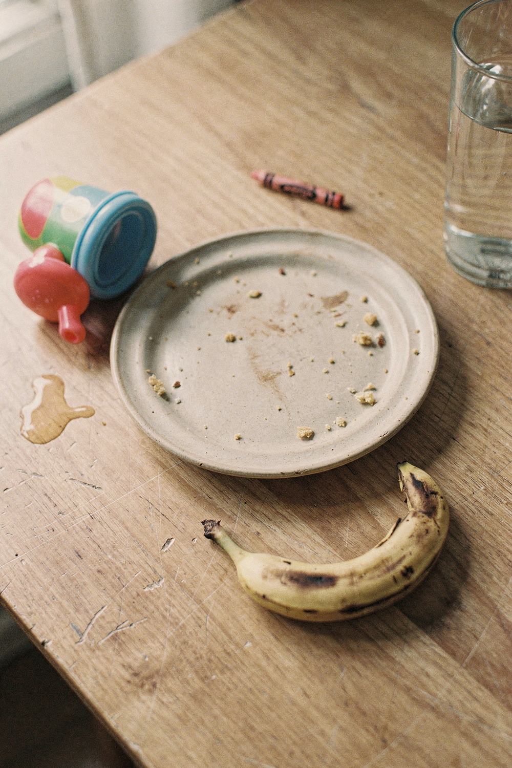 Empty plate with crumbs, a tipped sippy cup, a banana peel, and a glass of water on a dining table