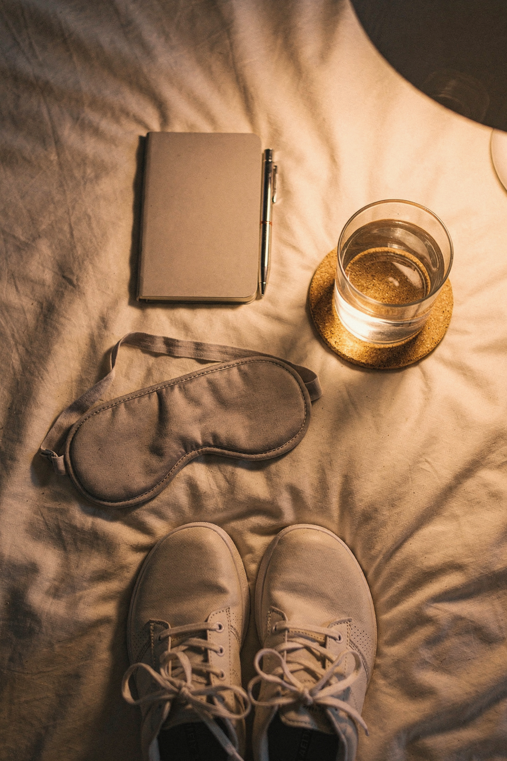 Sleep mask, journal with pen, a glass of water, and walking shoes on a cream bedspread