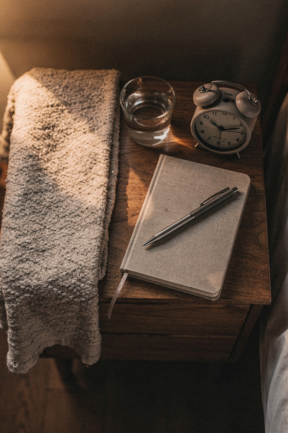 A folded blanket, a glass of water, a small alarm clock showing an early hour, and a closed journal with a pen on top. , no text, no logos