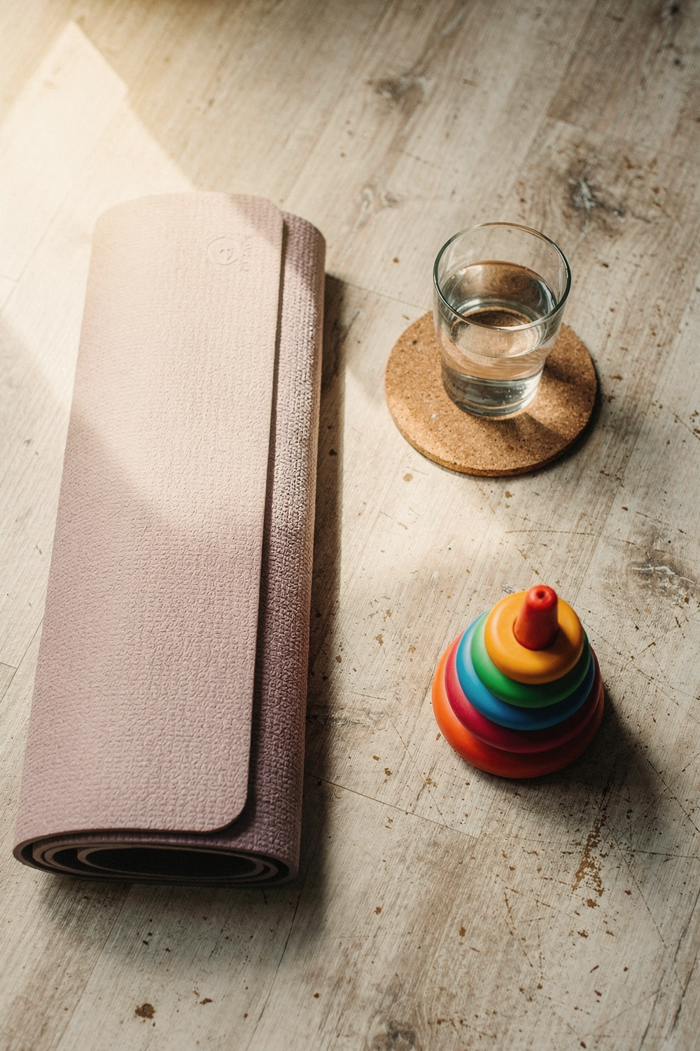 A folded yoga mat, a foam roller, and a glass of water on a cork coaster, with a child's stacking ring toy resting nearby. , no text