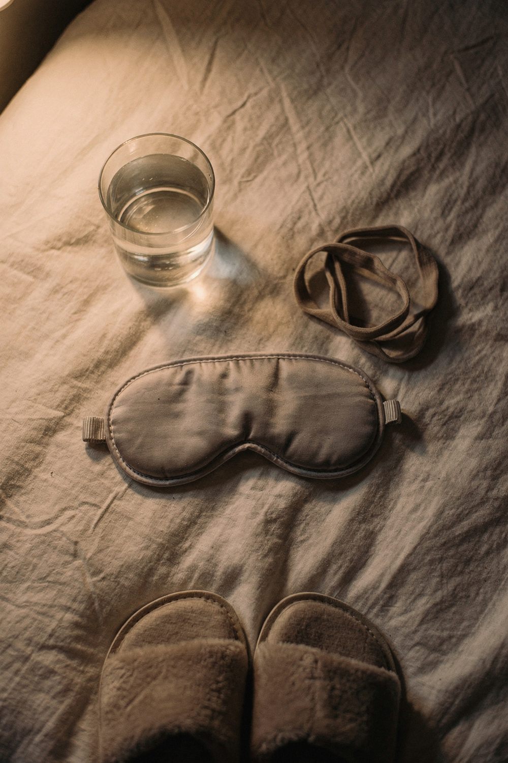 A sleep mask, a glass of water, a small stretching strap loosely coiled, and a pair of slippers at the edge of frame. , no text