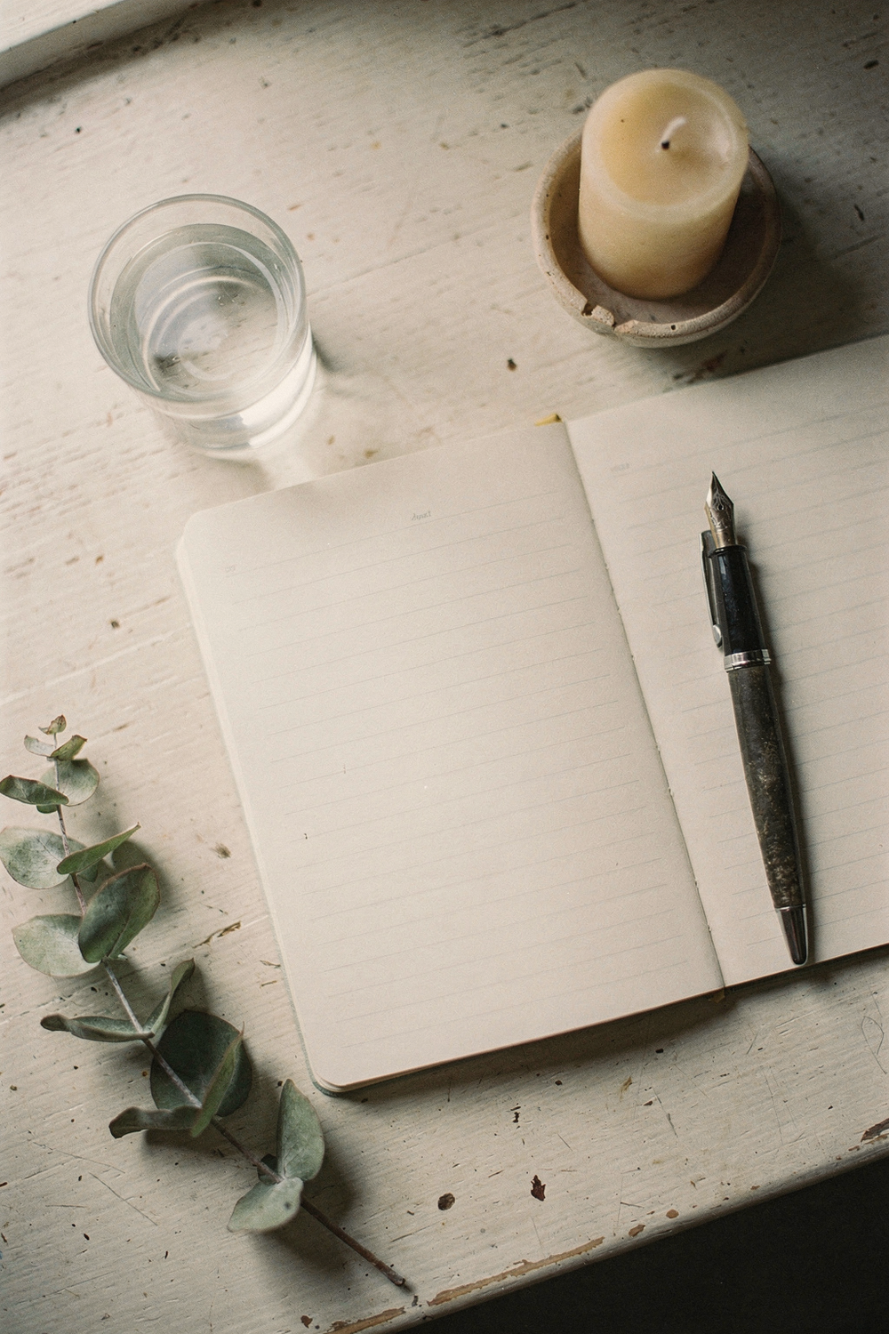A lined notebook open mid-page, a fountain pen, a small glass of water, a candle unlit, and a single eucalyptus stem. 50mm lens. , no text, no logos