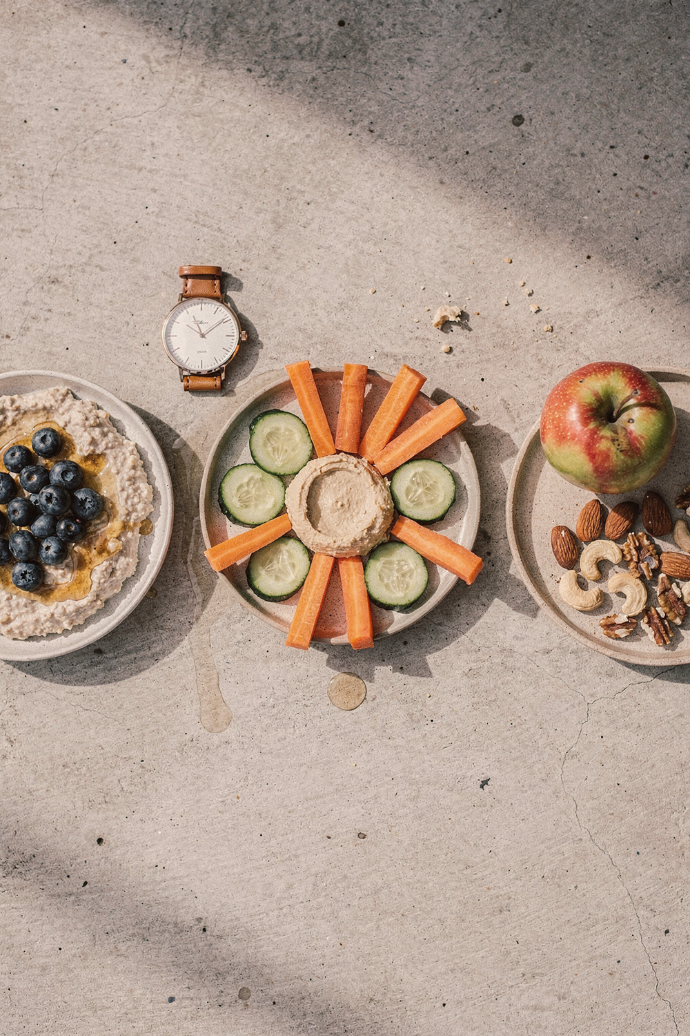 Three small plates arranged left to right: overnight oats, hummus with vegetables, and mixed nuts with an apple