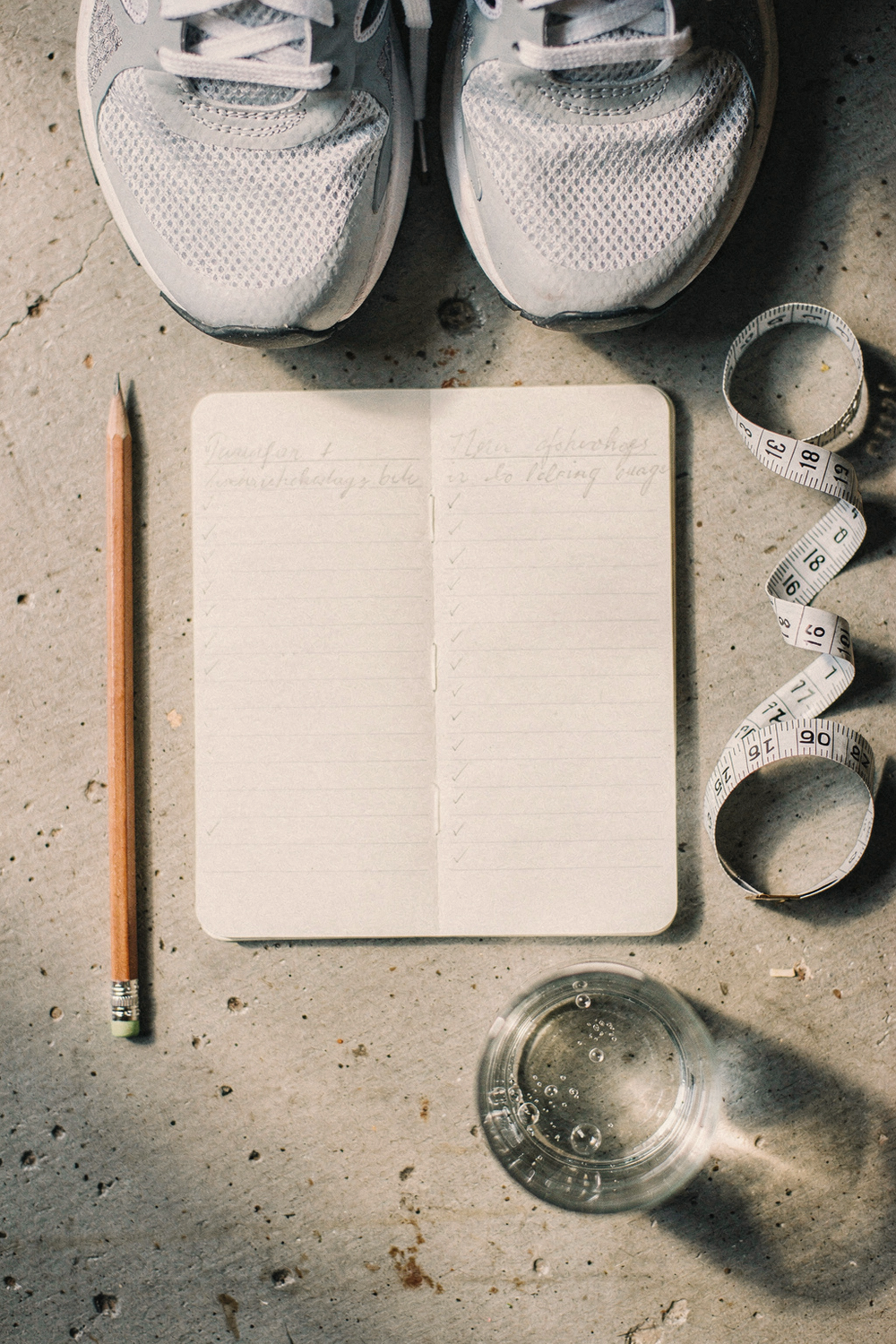 An open notebook with two handwritten columns and small checkmarks, a flexible tape measure laid in a gentle curve, a pencil, and a glass of water with condensation. 85mm lens, f/2. 8, Fuji Pro 400H tones. , no text