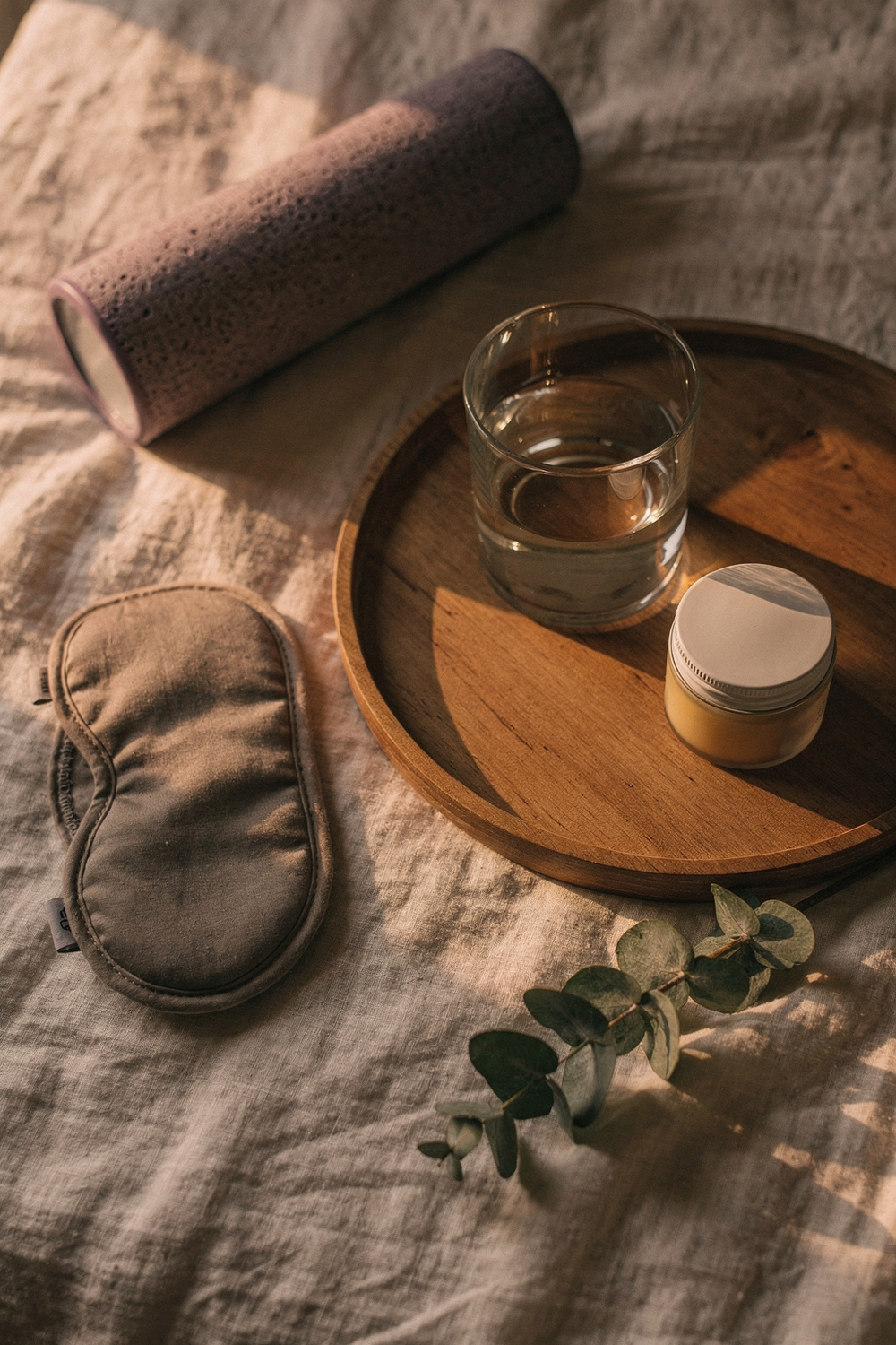 A folded sleep mask, a foam roller, a small jar of balm, and a glass of water on a wooden tray. A sprig of eucalyptus laid beside the tray. 85mm lens, f/2. 8, Kodak Portra 400 tones. , no text