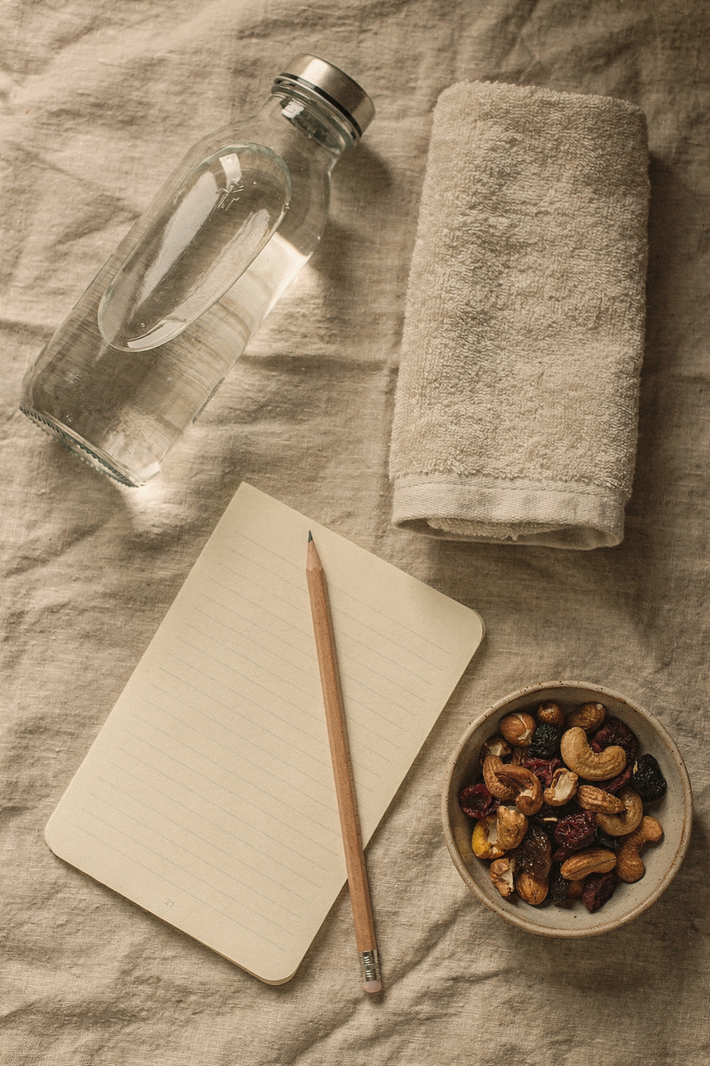 A glass water bottle, a folded towel, a simple lined notebook open with a pencil resting on it, and a small bowl of nuts and dried fruit. , no text, no logos
