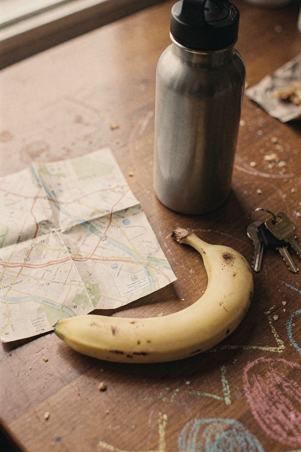 A folded park map, a reusable water bottle, a banana, a set of house keys, a kid's chalk drawing at the edge of the frame. , no text, no logos