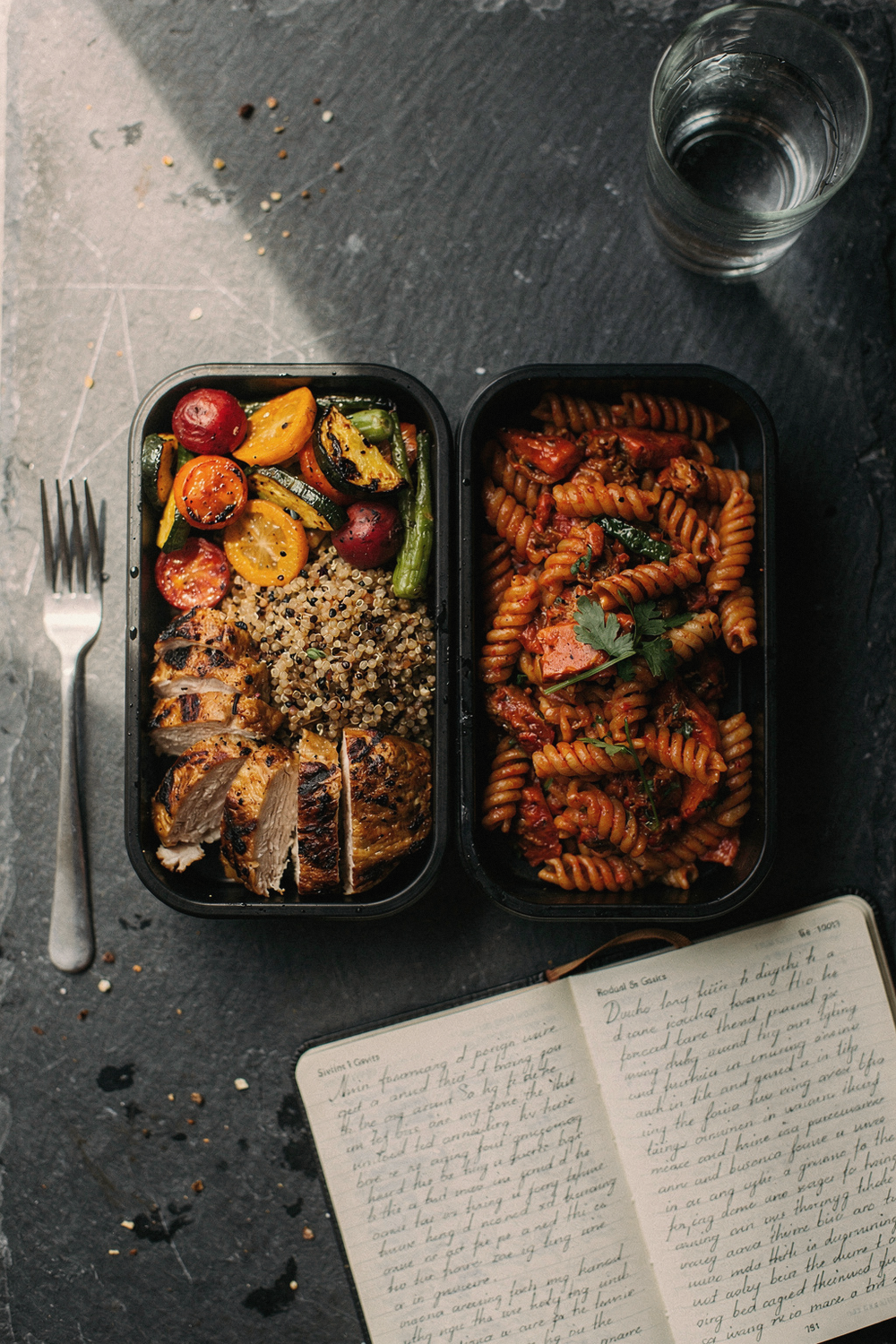 Two different meal prep containers side by side with different foods, a journal open to notes, a fork, and a water glass. , no faces, no hands