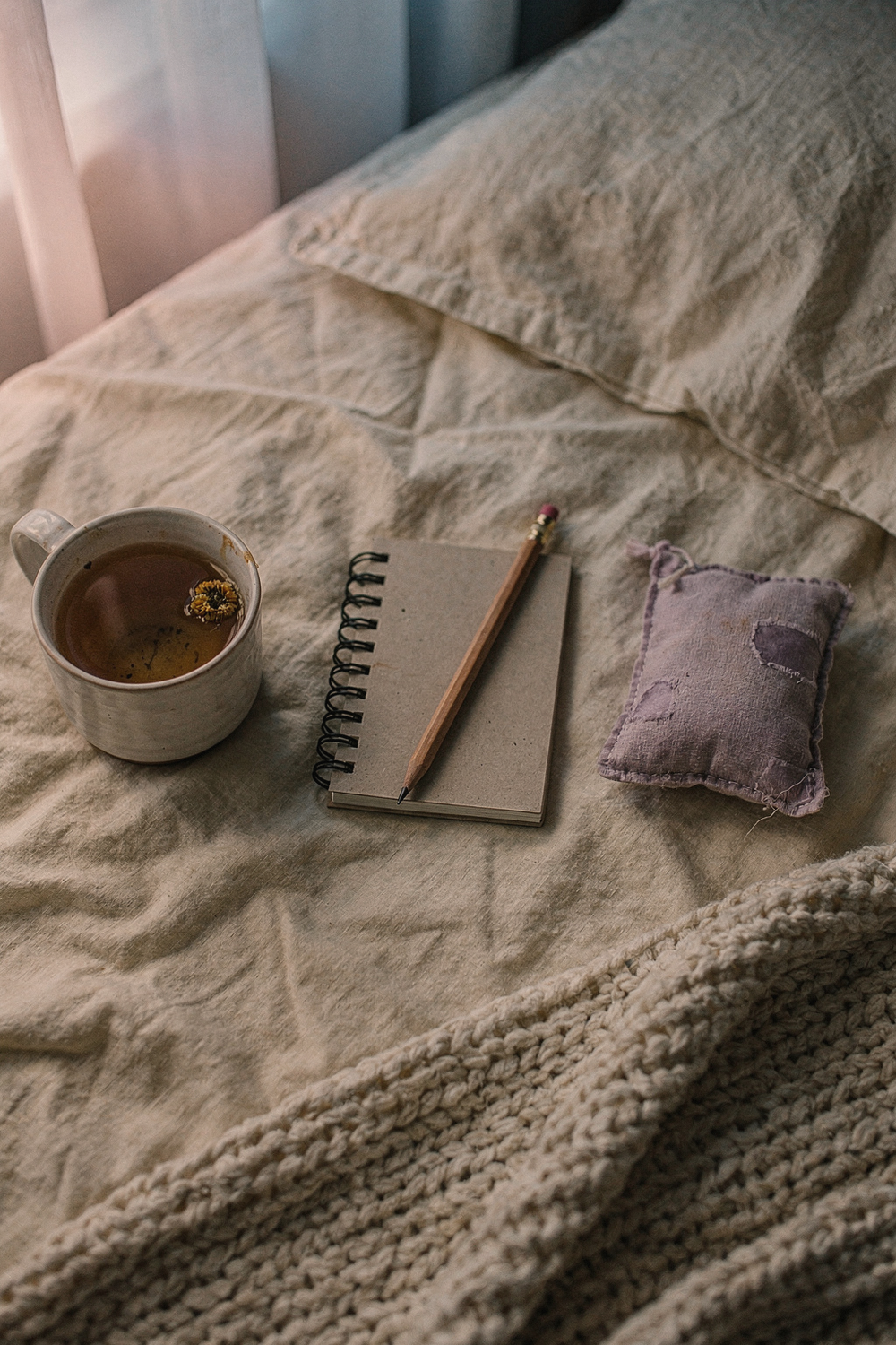 Pocket notebook with a pencil, a mug of herbal tea, a lavender sachet, and a knit blanket on linen sheets