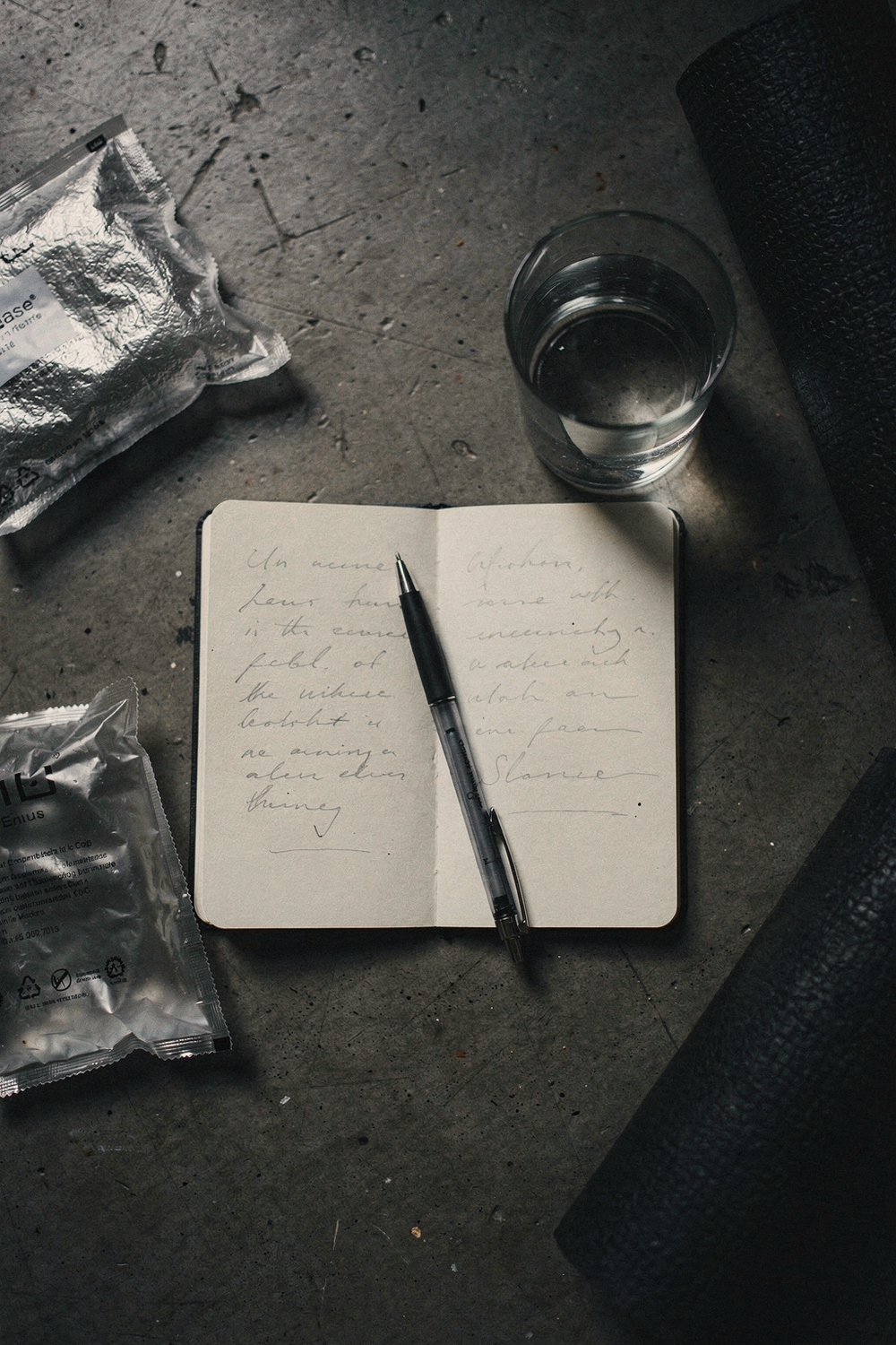 Unopened ice pack and foam roller at the edges, with a notebook, pen, and water glass in the center on concrete