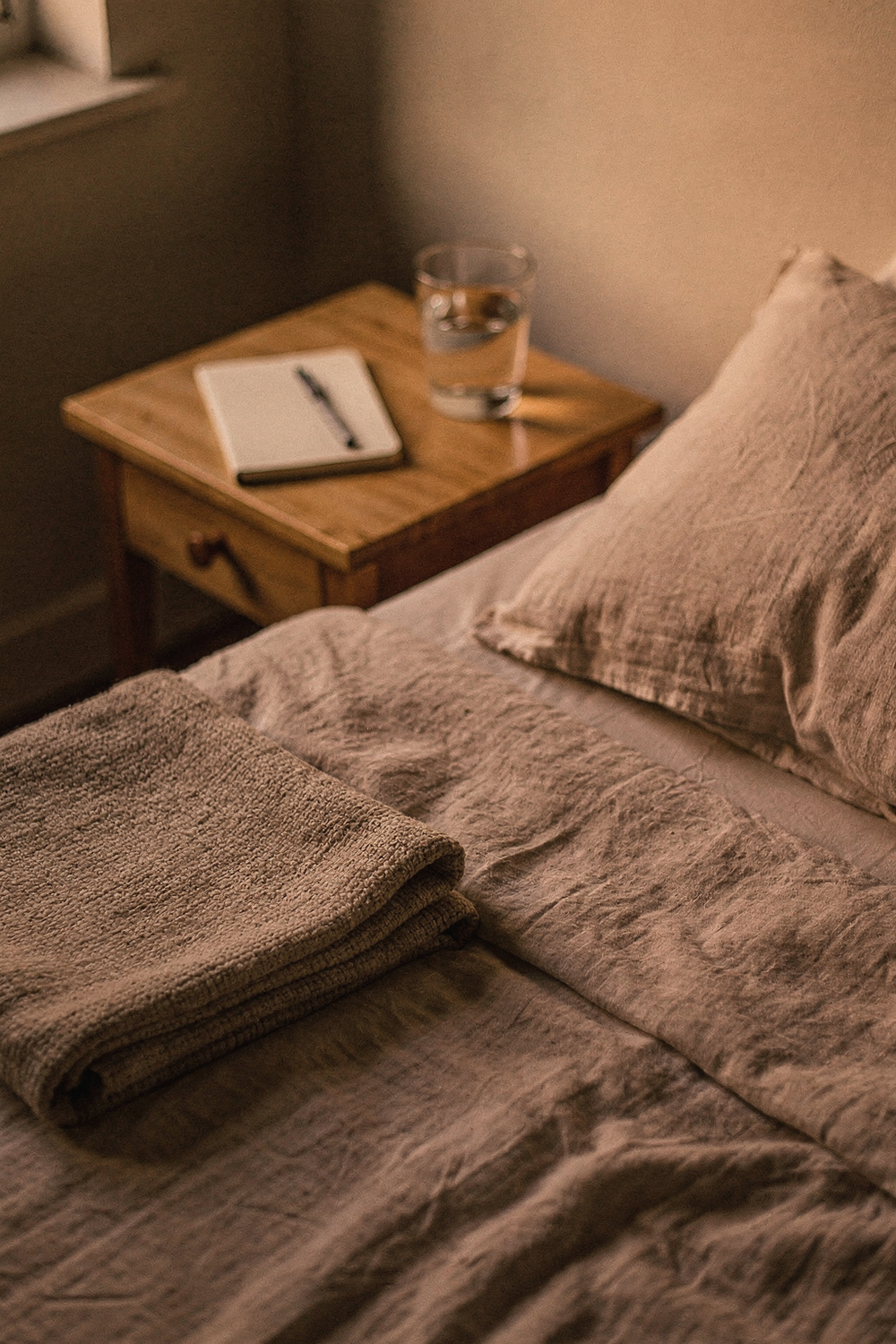 A neatly made bed with linen sheets, a folded throw, a glass of water and a small open notebook with a pen on the nightstand. NO faces, NO hands, NO text, NO logos