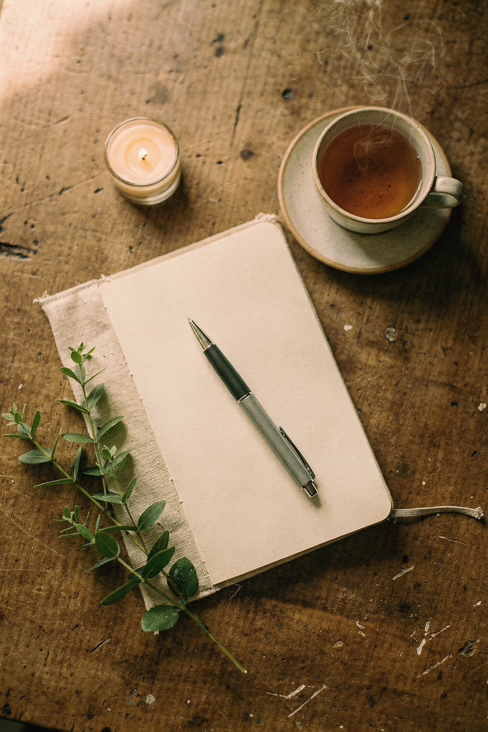 An open journal with a pen, a cup of tea, a small candle, and a sprig of greenery. NO faces, NO hands, NO text, NO logos