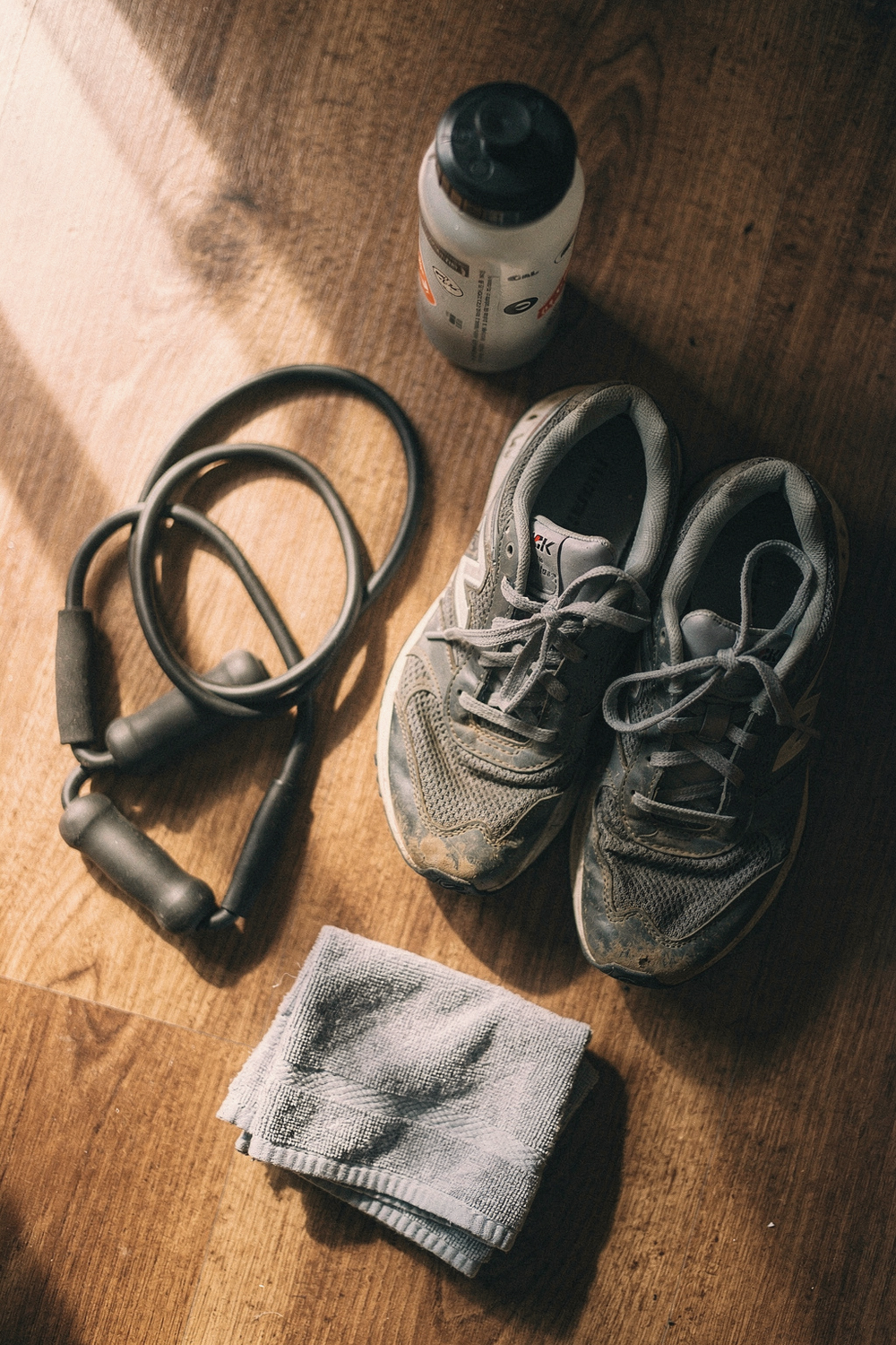 Worn sneakers, a resistance band, a water bottle with sticker residue, and a hand towel on a wood floor
