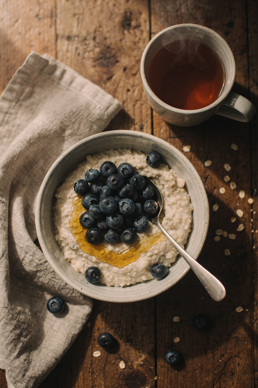 Oatmeal bowl topped with blueberries and honey, a spoon, and a cup of tea on a wooden table