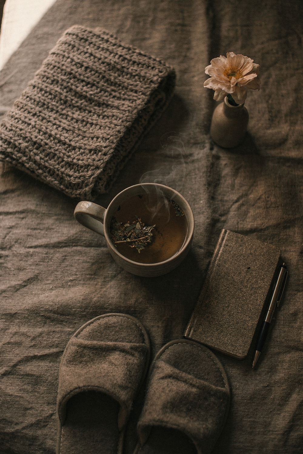 A cozy blanket folded, a cup of herbal tea, a small journal, a pair of slippers, and a single flower in a small vase. Restful, gentle