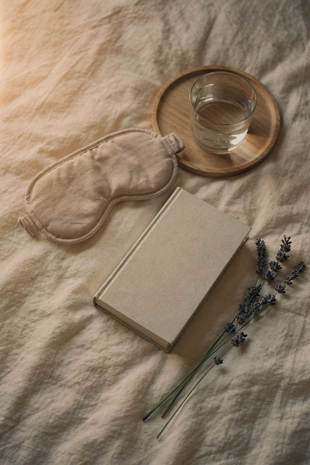 A folded sleep mask, a small glass of water on a wooden tray, a closed book, and a sprig of lavender. 85mm lens, f/2. 8, Fuji Pro 400H tones. , no text