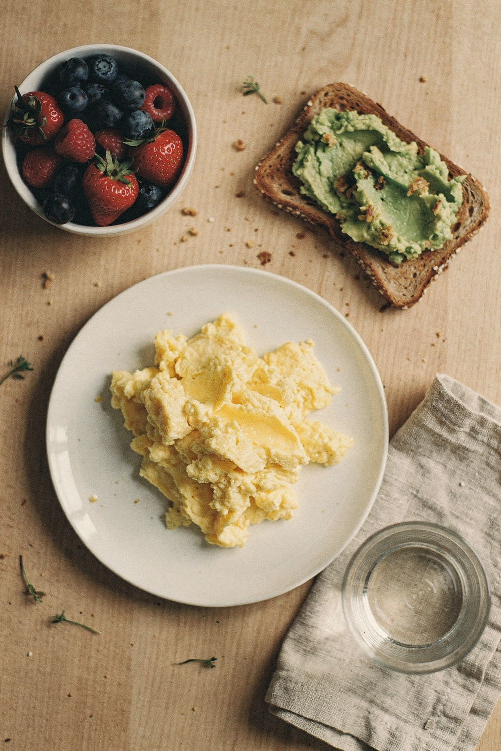A plate of scrambled eggs, a small bowl of berries, a slice of whole grain toast with avocado, and a glass of water on a linen napkin. , no text