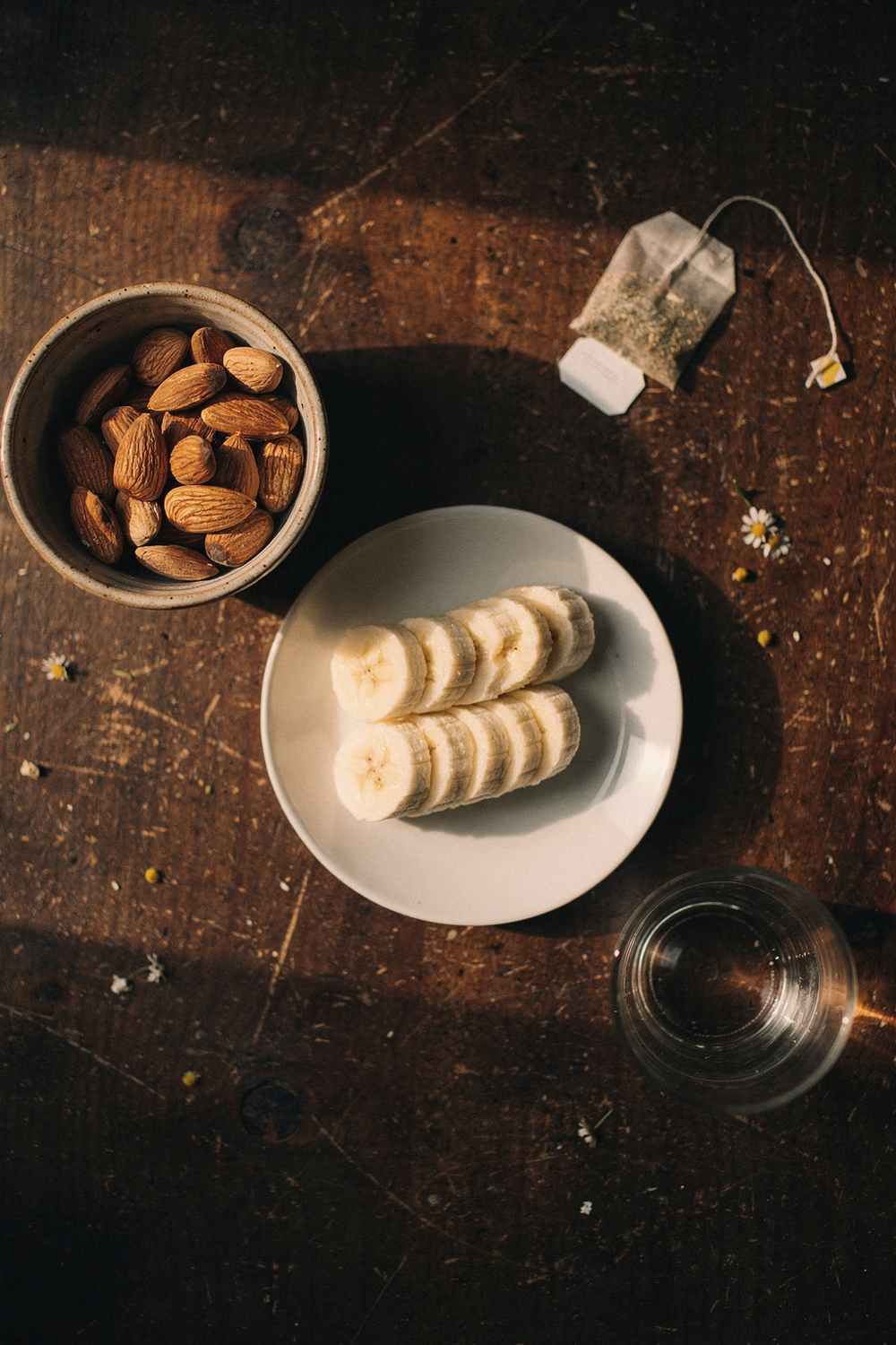 Small bowl of almonds, sliced banana on a plate, chamomile tea bag, and a glass of water