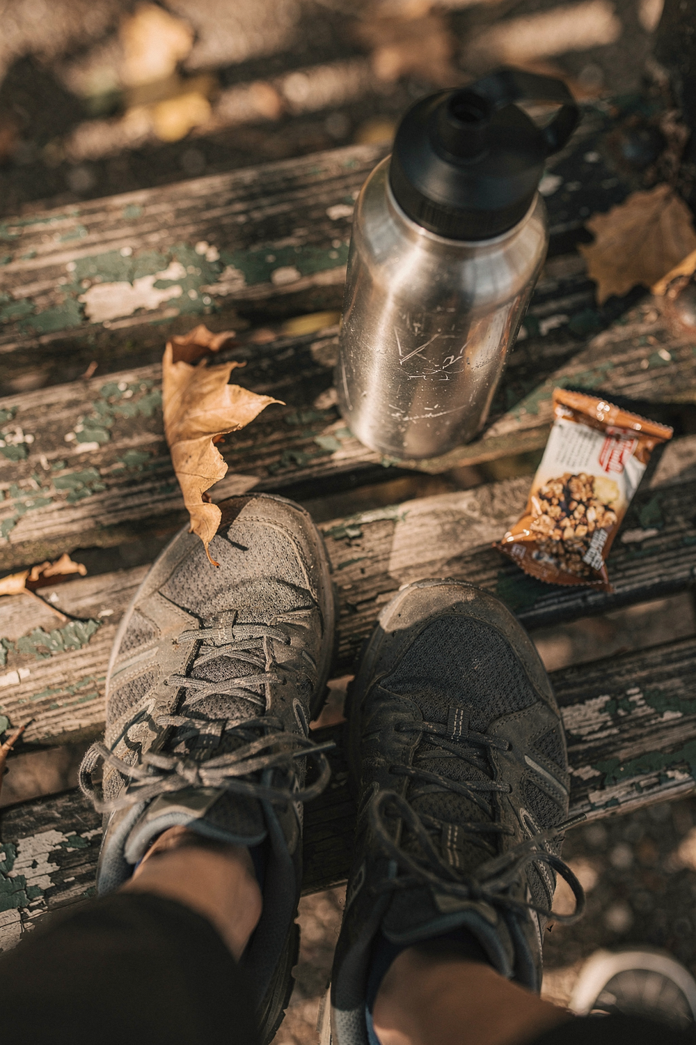Walking shoes, a scratched water bottle, a crumpled granola bar wrapper, and fallen leaves on a park bench