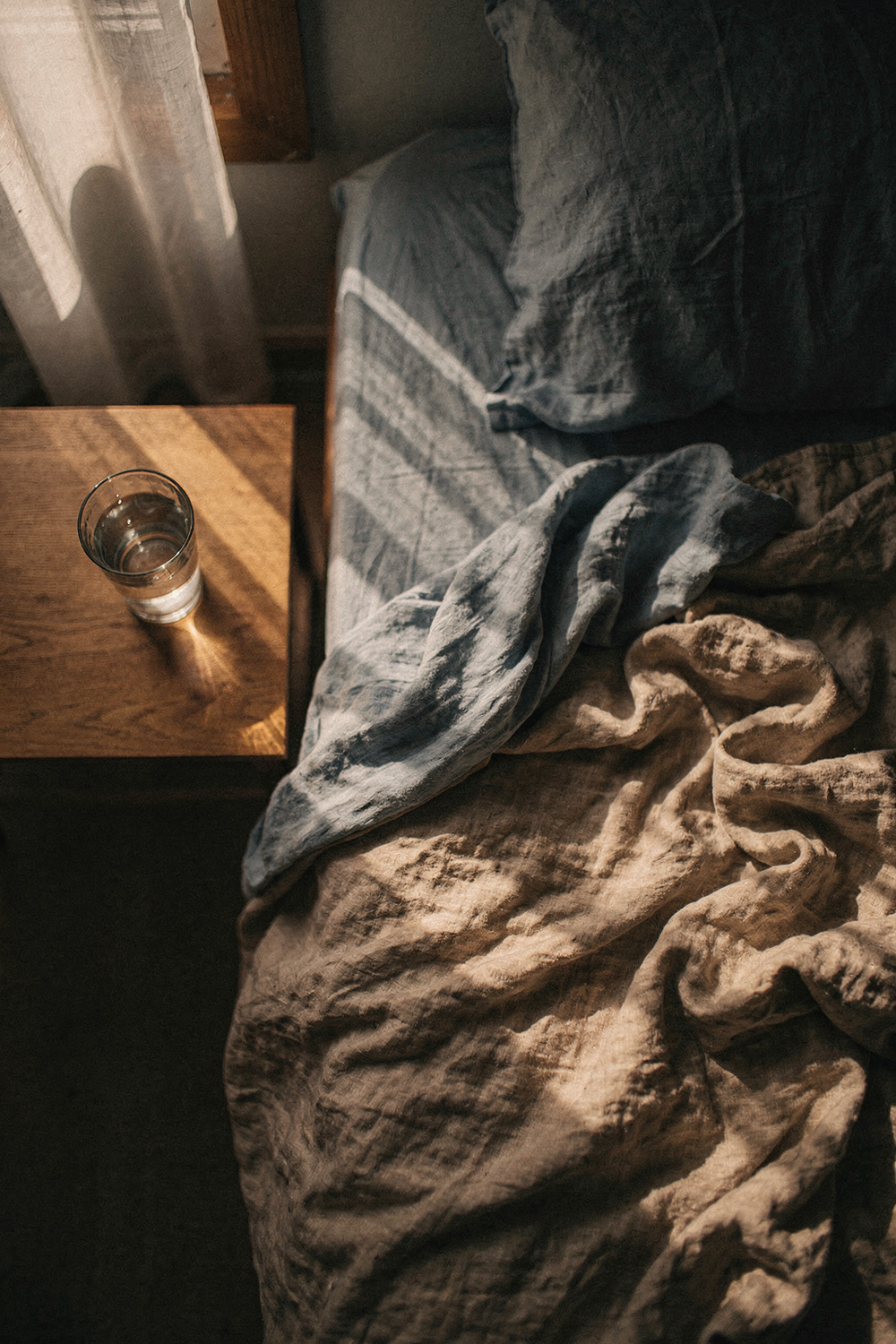 Rumpled linen sheets and a bedside table with a glass of water in soft dawn light