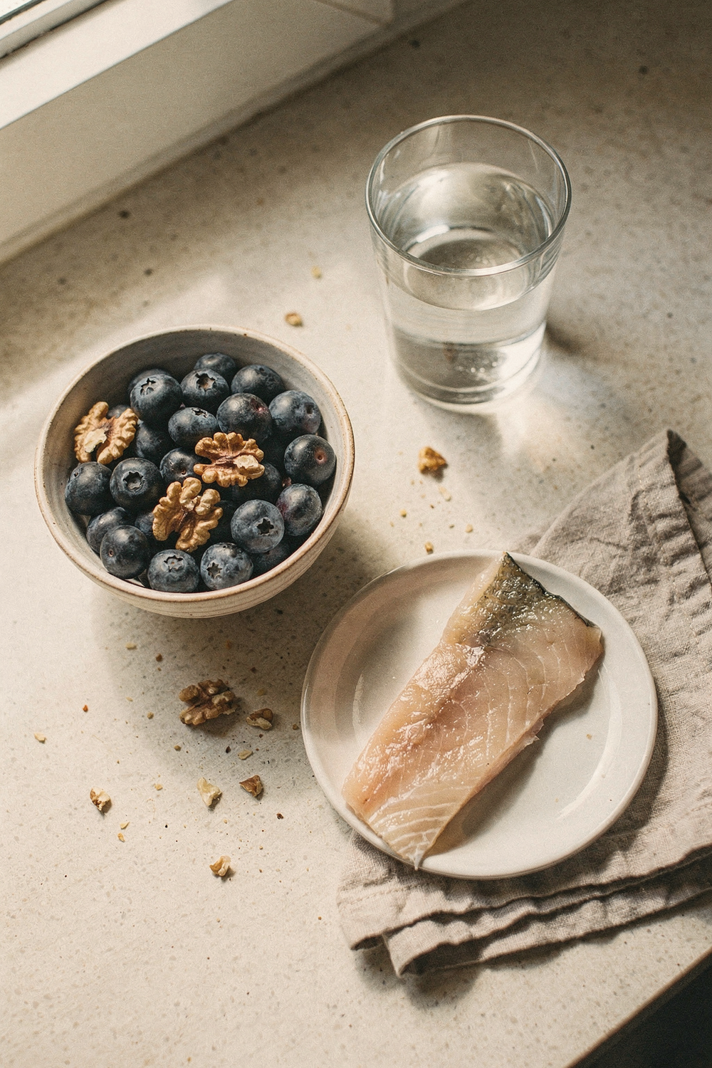 A bowl of blueberries and walnuts, a glass of water, a piece of fatty fish on a small plate, and a folded napkin. Fresh, grounded, honest. , no text, no logos