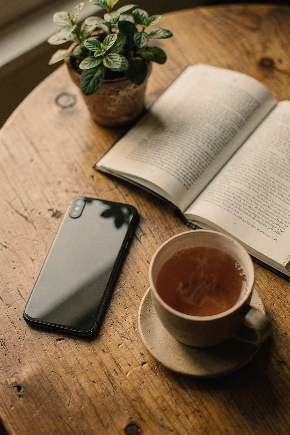 Phone face-down next to a cup of tea, an open book, and a small potted plant
