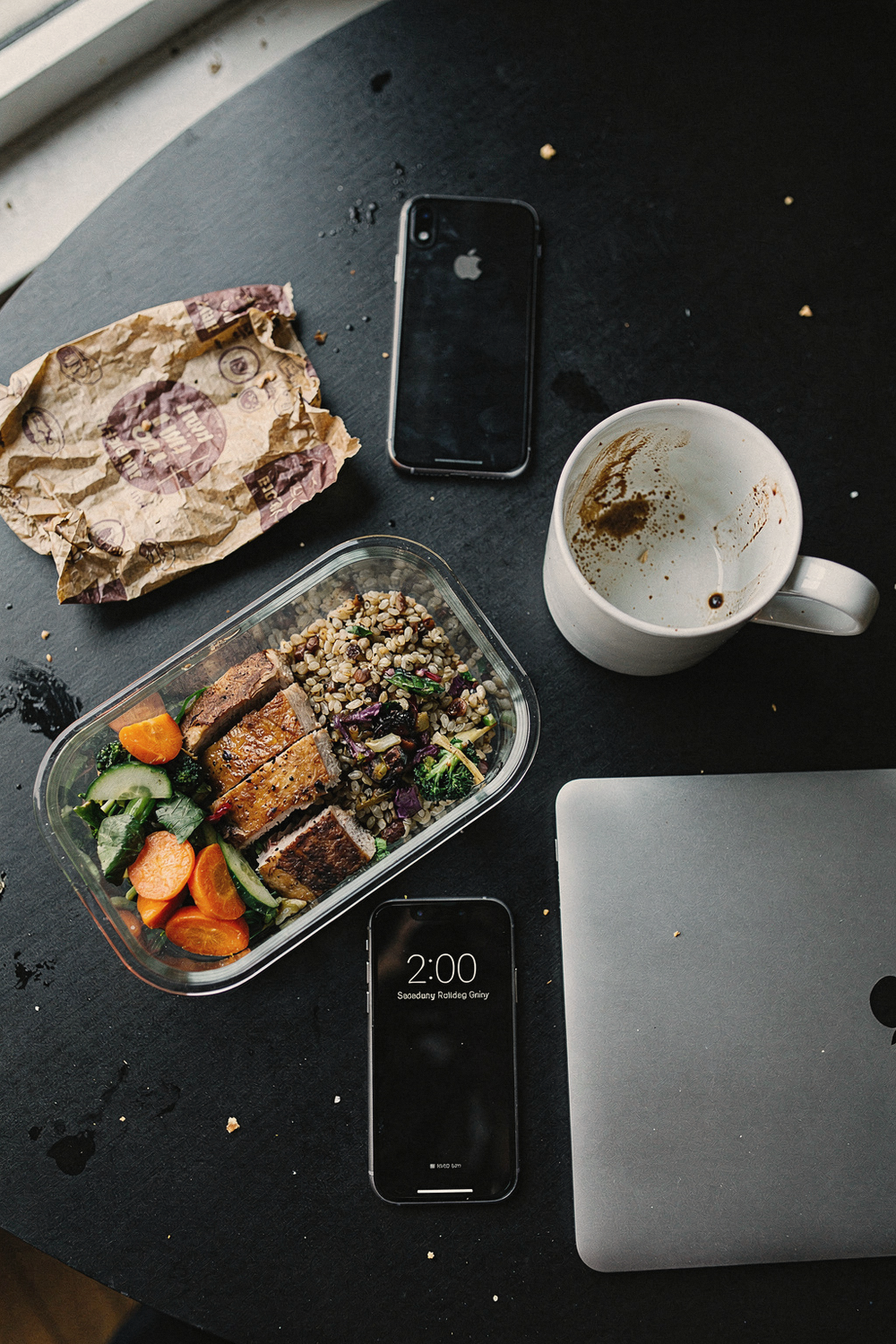 An energy-crashing fast food wrapper crumpled beside a balanced meal in a glass container, a coffee cup half empty, a phone showing 2:00pm, and a closed laptop. Honest, relatable