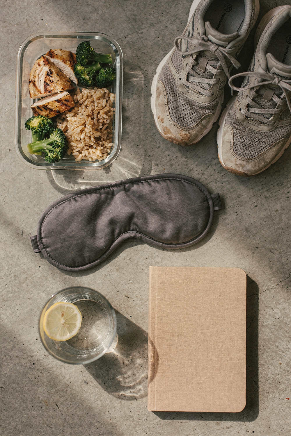 A sleep mask, a meal prep container, running shoes, a small journal, and a glass of water with lemon. Grounded, systematic